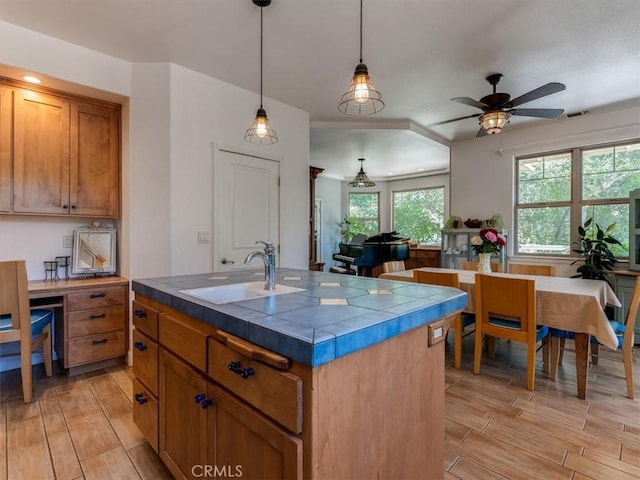 kitchen featuring tile countertops, sink, ceiling fan, an island with sink, and decorative light fixtures