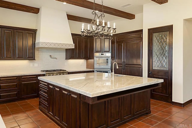 kitchen featuring premium range hood, a kitchen island with sink, decorative light fixtures, beam ceiling, and a chandelier