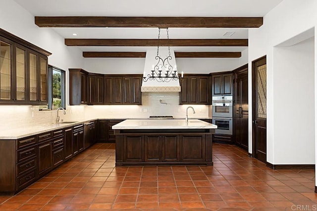 kitchen featuring backsplash, double oven, beam ceiling, hanging light fixtures, and an island with sink