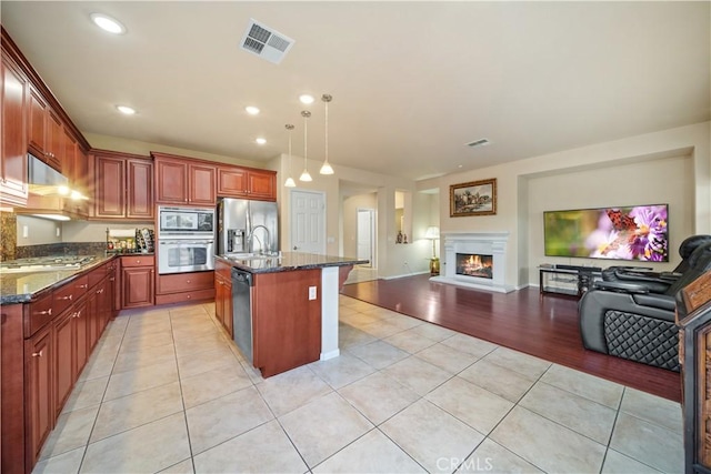 kitchen featuring light tile patterned floors, appliances with stainless steel finishes, a kitchen island with sink, dark stone countertops, and pendant lighting