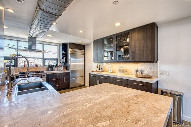 kitchen featuring decorative backsplash, light stone countertops, dark brown cabinetry, built in fridge, and range hood