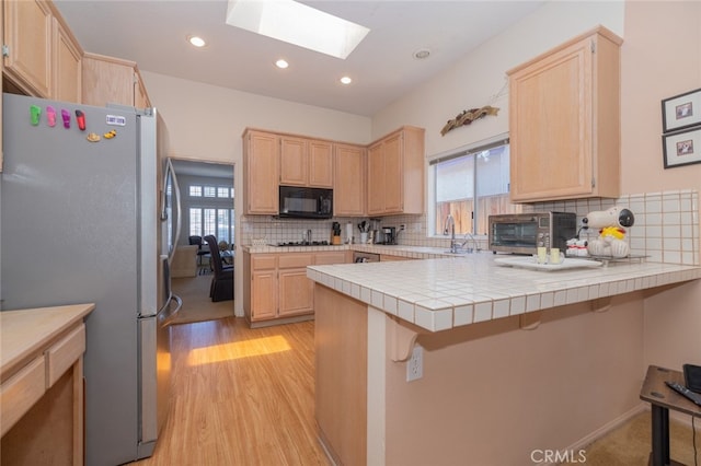 kitchen with tile counters, a skylight, kitchen peninsula, stainless steel refrigerator, and a breakfast bar area