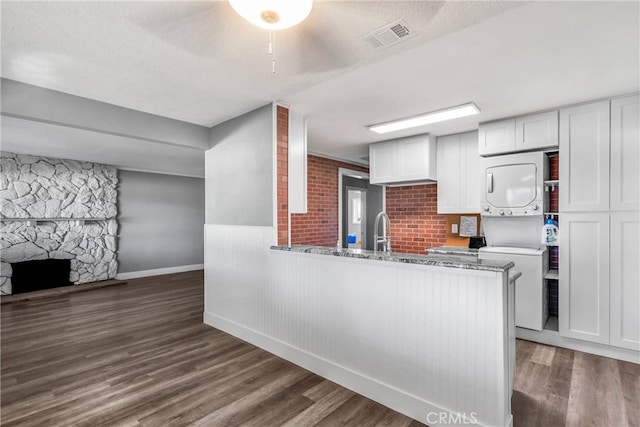 kitchen featuring visible vents, wood finished floors, stacked washing maching and dryer, a stone fireplace, and white cabinets