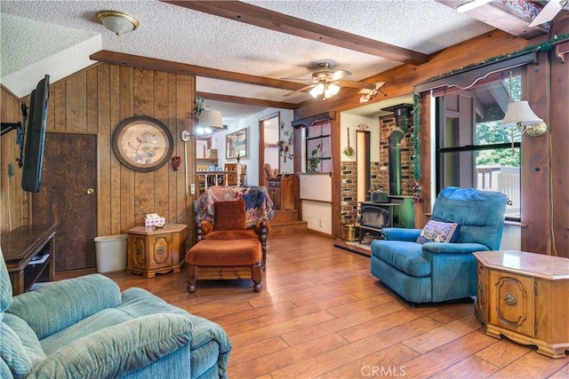 living room with vaulted ceiling with beams, a textured ceiling, a wood stove, wooden walls, and light hardwood / wood-style floors