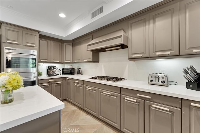 kitchen with gray cabinetry, ornamental molding, a raised ceiling, custom exhaust hood, and appliances with stainless steel finishes