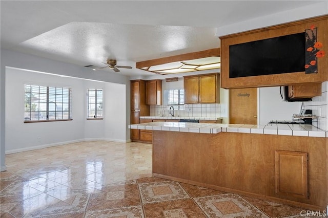 kitchen with tile countertops, ceiling fan, a peninsula, backsplash, and brown cabinetry