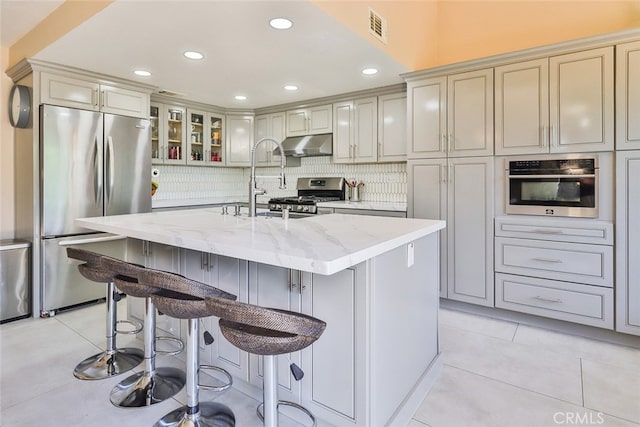 kitchen featuring light tile patterned flooring, backsplash, a kitchen island with sink, light stone countertops, and appliances with stainless steel finishes