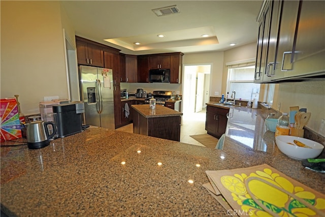 kitchen featuring stainless steel refrigerator with ice dispenser, range, dark stone countertops, a raised ceiling, and kitchen peninsula