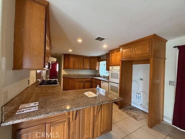 24062 Eucalyptus Ave, Moreno Valley, CA 92553 - kitchen with visible vents, brown cabinetry, a peninsula, white appliances, and light stone countertops