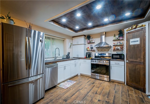 kitchen with white cabinets, a raised ceiling, sink, wall chimney range hood, and appliances with stainless steel finishes