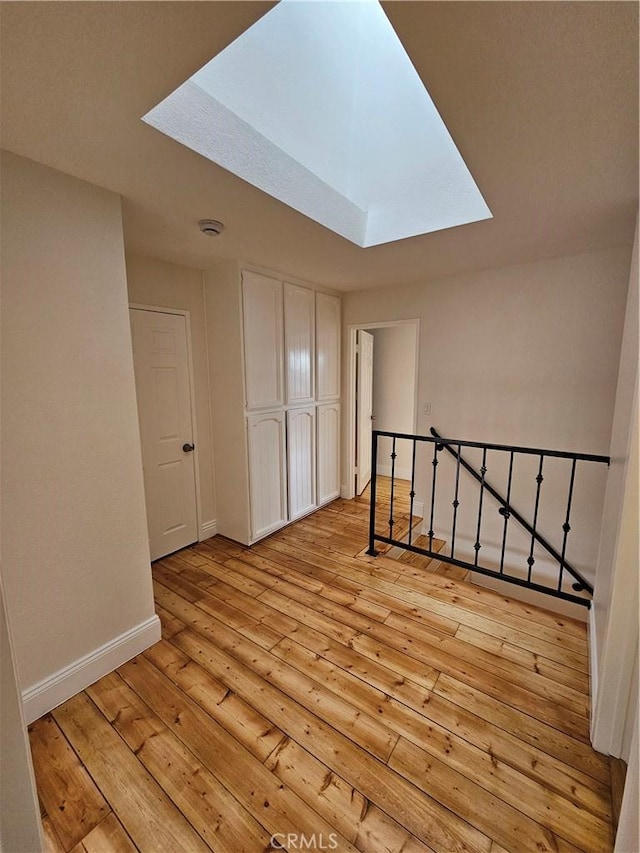 corridor featuring baseboards, an upstairs landing, light wood-style floors, and a skylight