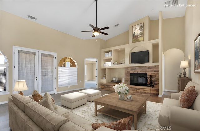 living room featuring ceiling fan, a high ceiling, built in shelves, a stone fireplace, and french doors