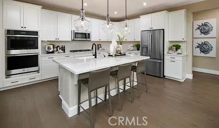 kitchen with white cabinetry, dark wood-type flooring, stainless steel appliances, decorative light fixtures, and a kitchen island with sink