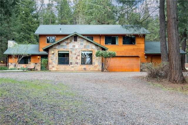 chalet / cabin featuring stone siding, roof with shingles, a chimney, gravel driveway, and a garage