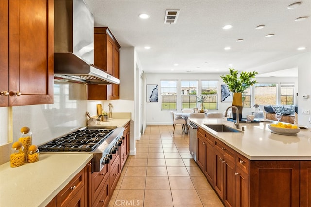 kitchen featuring stainless steel appliances, sink, wall chimney range hood, light tile patterned floors, and a center island with sink