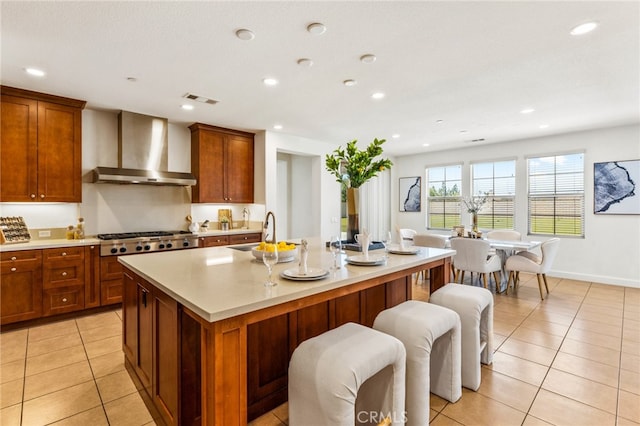 kitchen with stainless steel gas stovetop, wall chimney range hood, sink, an island with sink, and light tile patterned flooring