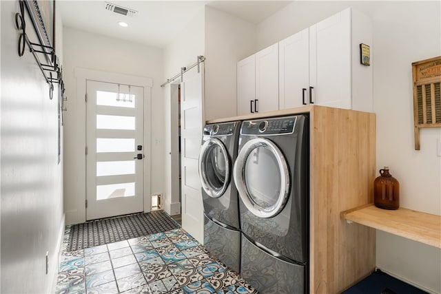 clothes washing area with a barn door, washer and dryer, light tile patterned floors, and cabinets