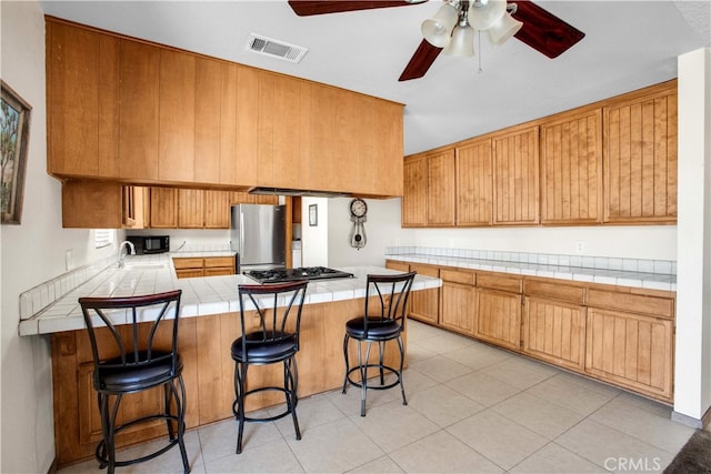 kitchen with appliances with stainless steel finishes, a breakfast bar, kitchen peninsula, tile countertops, and ceiling fan