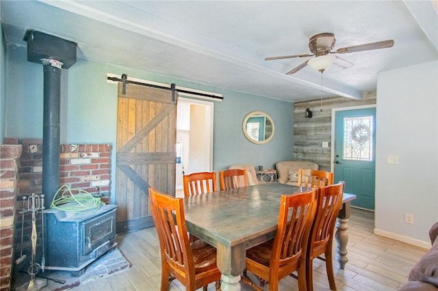 dining room with beam ceiling, a ceiling fan, a barn door, wood finished floors, and a wood stove