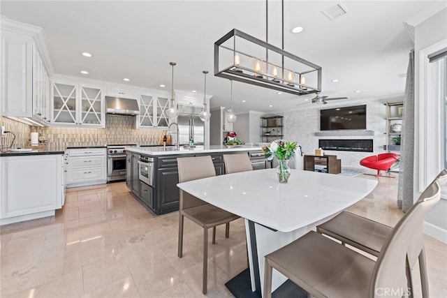 kitchen featuring decorative backsplash, a center island with sink, white cabinetry, pendant lighting, and ventilation hood