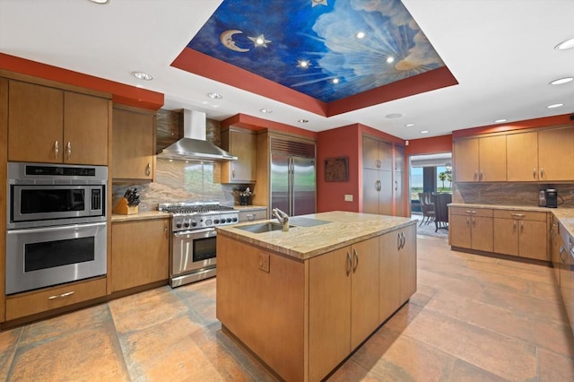 kitchen featuring high end appliances, a center island with sink, a raised ceiling, wall chimney range hood, and decorative backsplash