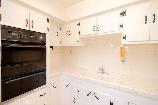 kitchen with a sink, tile counters, white cabinetry, dobule oven black, and tasteful backsplash