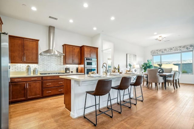 kitchen featuring appliances with stainless steel finishes, wall chimney exhaust hood, a breakfast bar, a kitchen island with sink, and light hardwood / wood-style floors