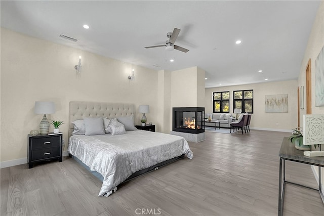 bedroom featuring a multi sided fireplace, light hardwood / wood-style flooring, and ceiling fan