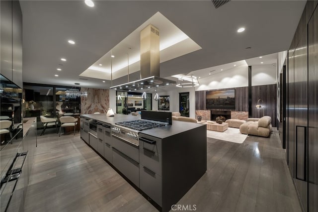 kitchen with a stone fireplace, island exhaust hood, a spacious island, stainless steel gas stovetop, and a tray ceiling