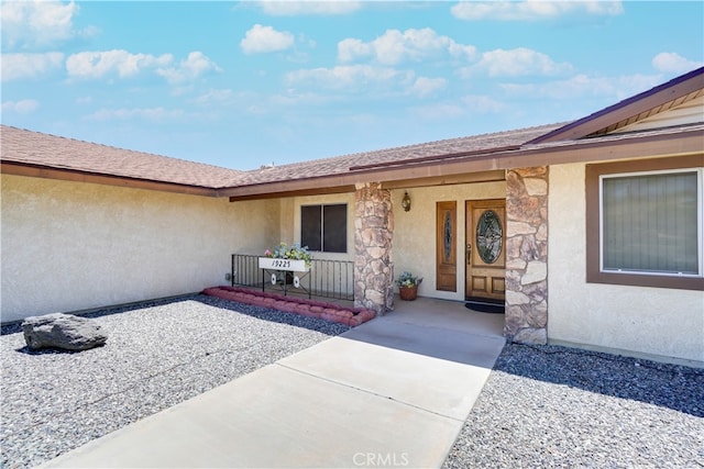 view of exterior entry with stone siding, a shingled roof, and stucco siding