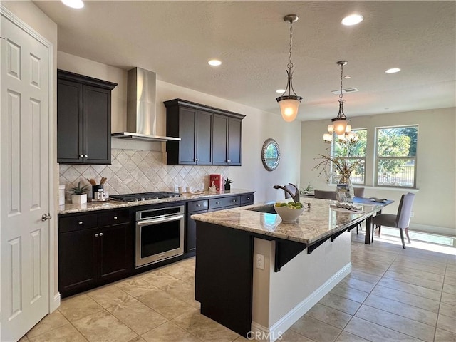 kitchen with pendant lighting, sink, appliances with stainless steel finishes, a kitchen island with sink, and wall chimney exhaust hood