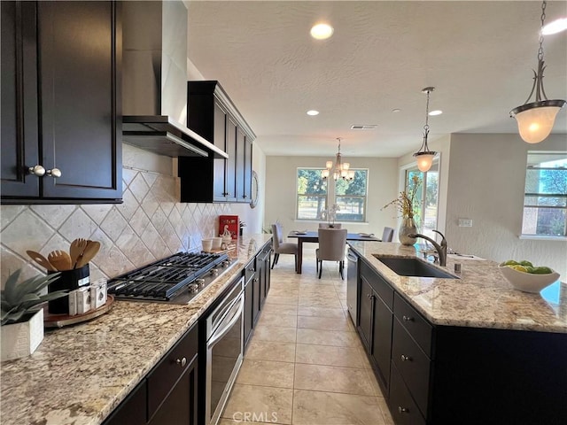 kitchen with wall chimney range hood, sink, appliances with stainless steel finishes, light stone counters, and decorative light fixtures