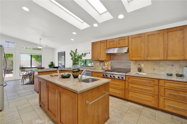 kitchen featuring light stone countertops, backsplash, vaulted ceiling with skylight, stainless steel appliances, and an island with sink