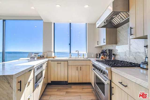 kitchen featuring stainless steel appliances, wall chimney exhaust hood, a water view, light hardwood / wood-style flooring, and light brown cabinets