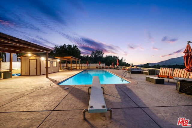 pool at dusk featuring a patio area, an outdoor hangout area, a storage unit, a diving board, and a mountain view