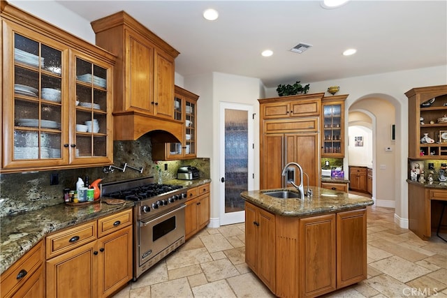 kitchen with light tile patterned flooring, high end stove, sink, decorative backsplash, and a center island with sink