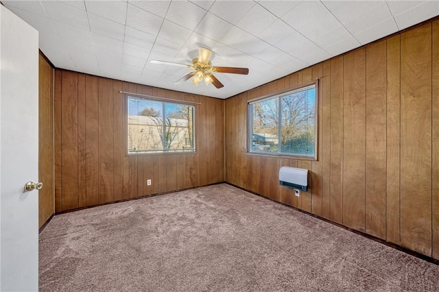 carpeted spare room featuring heating unit, wood walls, ceiling fan, and a healthy amount of sunlight