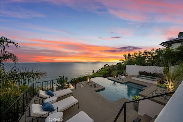 pool at dusk with an in ground hot tub, a water view, and a patio area