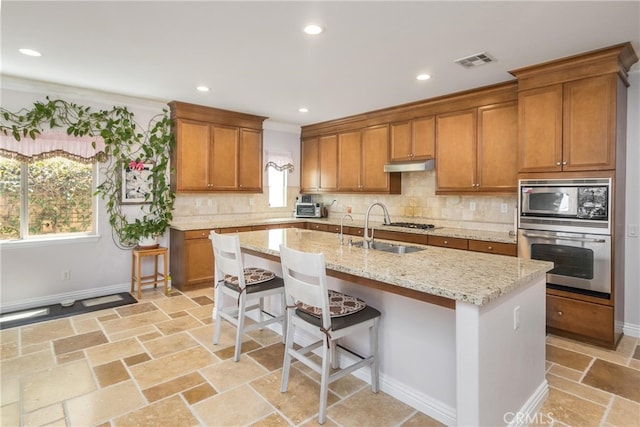 kitchen with a center island with sink, sink, appliances with stainless steel finishes, tasteful backsplash, and light stone counters