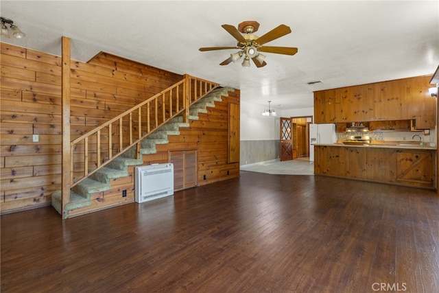 unfurnished living room with heating unit, ceiling fan with notable chandelier, wood walls, and dark wood-type flooring