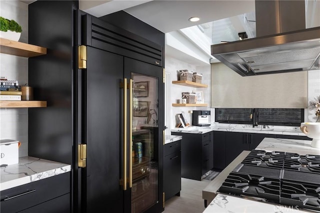 kitchen with backsplash, light stone counters, black gas stovetop, stainless steel built in refrigerator, and exhaust hood