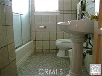 bathroom featuring tile walls, bath / shower combo with glass door, and toilet