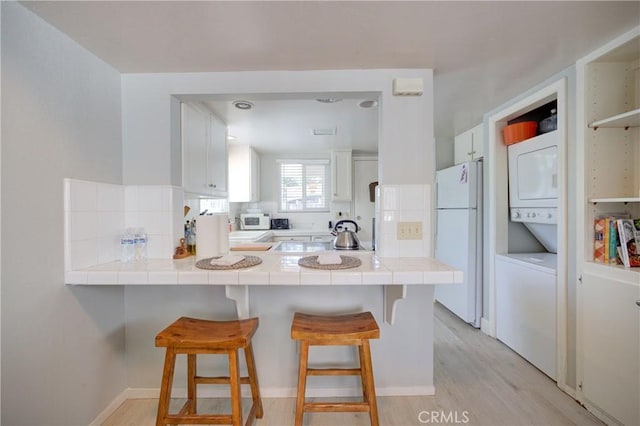 kitchen featuring stacked washing maching and dryer, a breakfast bar, white cabinets, tile counters, and white appliances