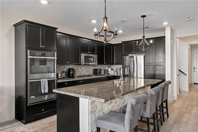 kitchen with hanging light fixtures, an island with sink, dark stone counters, light wood-type flooring, and stainless steel appliances