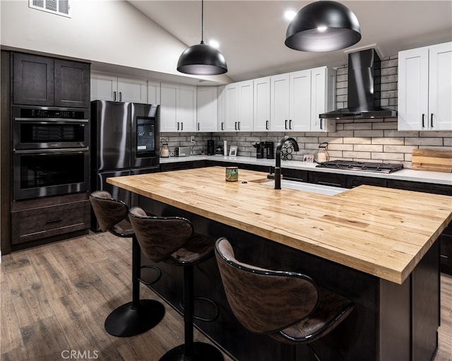 kitchen featuring wall chimney range hood, a kitchen island with sink, pendant lighting, wooden counters, and stainless steel appliances