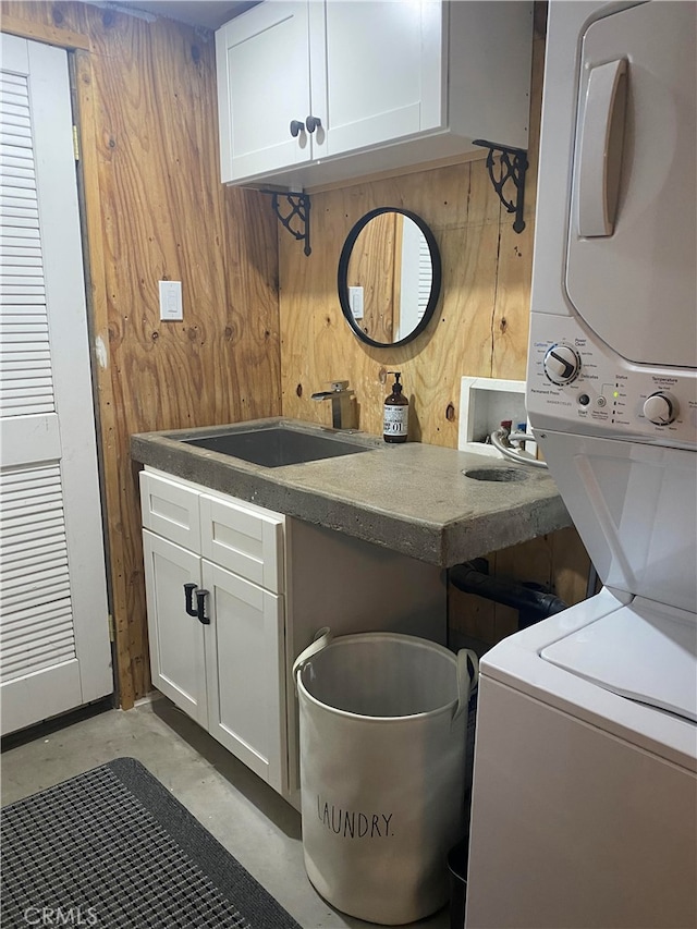 clothes washing area with stacked washer and clothes dryer, wood walls, sink, and cabinets