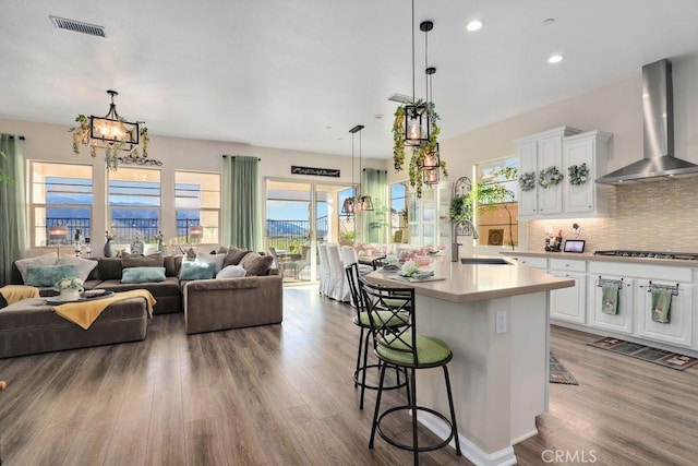 kitchen with a kitchen island with sink, hanging light fixtures, wall chimney exhaust hood, a kitchen bar, and white cabinetry