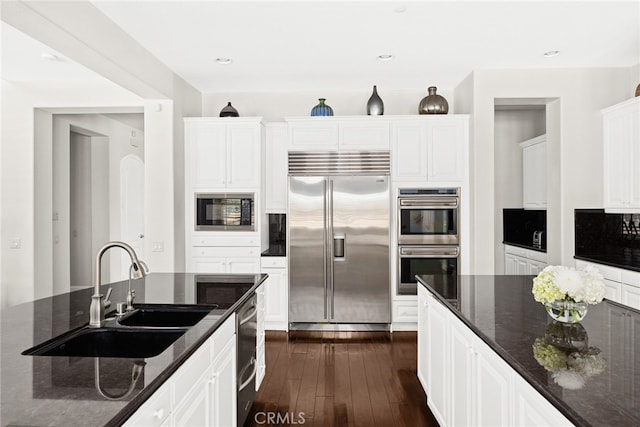 kitchen with dark stone countertops, white cabinets, built in appliances, dark hardwood / wood-style flooring, and sink