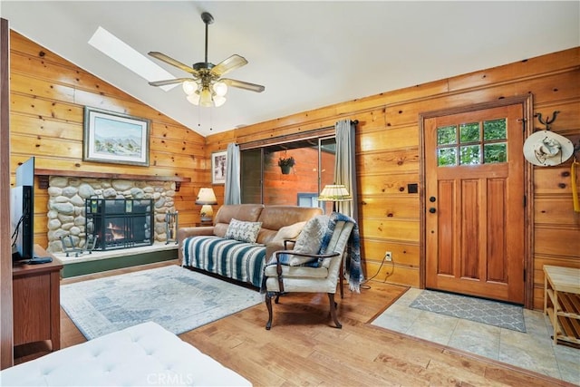 living room featuring ceiling fan, wooden walls, a fireplace, vaulted ceiling with skylight, and light hardwood / wood-style flooring