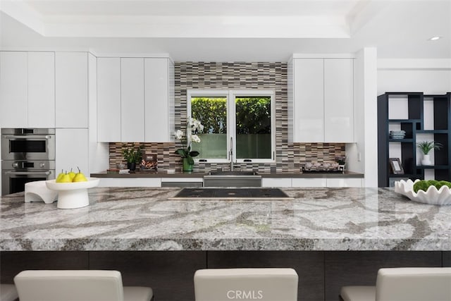 kitchen with white cabinetry, backsplash, light stone countertops, black electric stovetop, and double oven